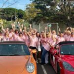 Participantes en polos roses derrière des Porsche orange et rouge au Rallye des Demoiselles.