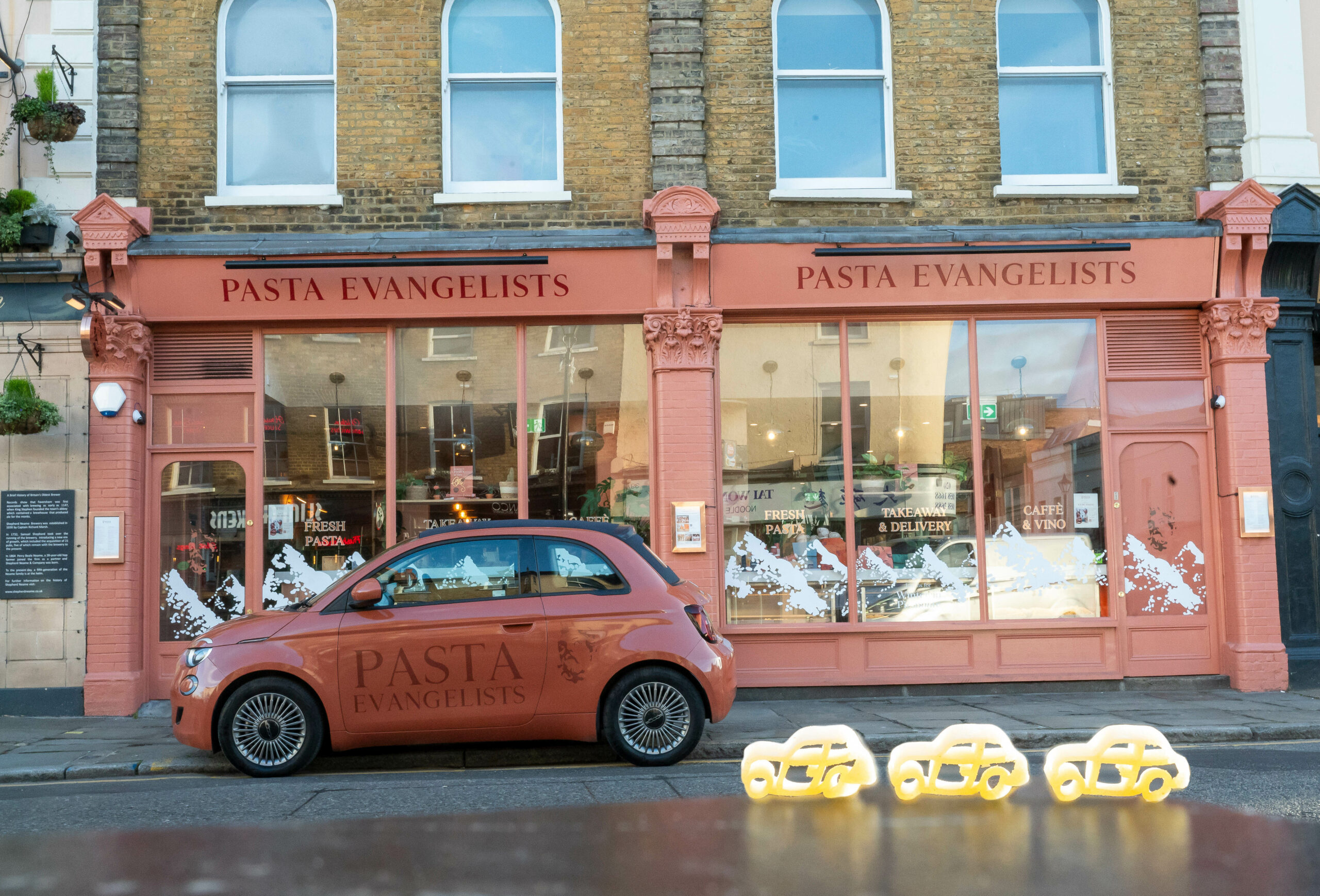 Fiat 500 aux couleurs de Pasta Evangelists devant une boutique, avec des pâtes en forme de voiture au premier plan